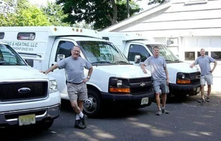 Three men pose with work vehicles in front of a building.