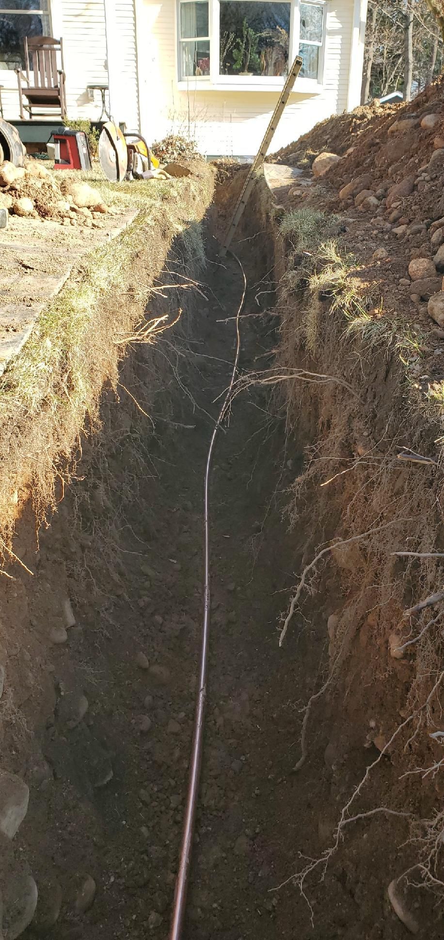 A long, narrow trench with a copper pipe running down the center, near a house.