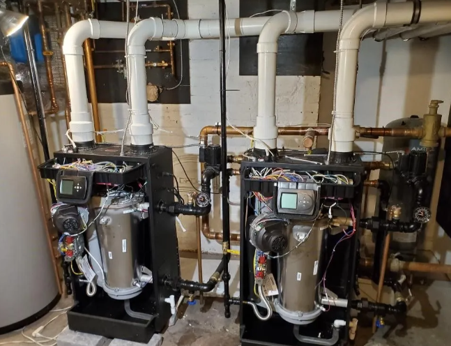 Two black and silver boilers with white pipes, installed in a basement, connected by copper piping.