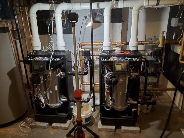 Two black, industrial boilers with white pipes in a basement.
