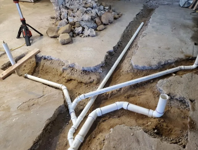 White PVC pipes installed in a trench dug into a concrete floor, with dirt and rocks visible.