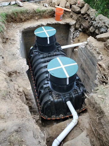 Black septic tank in a trench, two green lids, white pipes, dirt around.