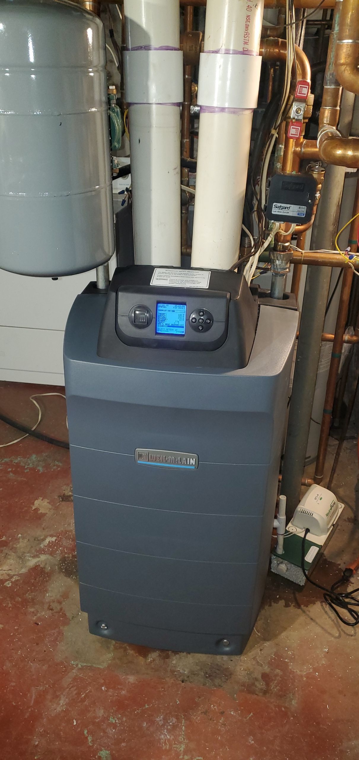 A gray water softener unit with white pipes and a blue display, installed in a basement.