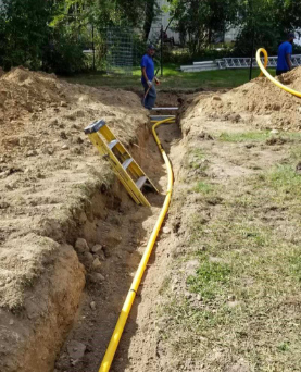 Construction workers installing yellow gas line in a trench in a backyard; a ladder is present.