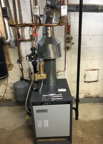 A boiler with a chimney in a basement. Copper pipes and a black pipe are visible on the wall.