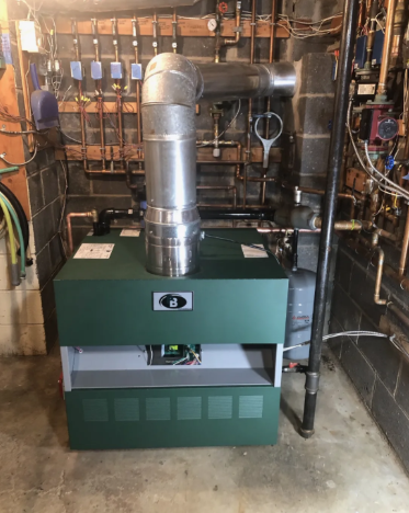 Green boiler in a utility room with silver vent pipe, surrounded by pipes and wiring.