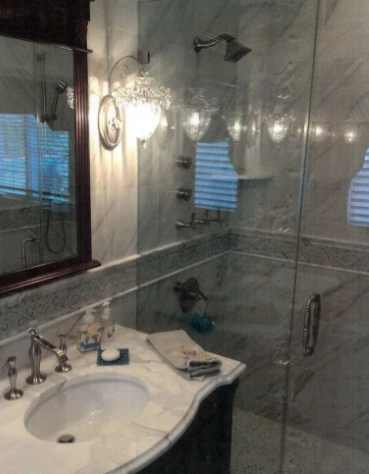 Bathroom with marble walls and countertop, glass shower, ornate light fixture, and sink with chrome fixtures.