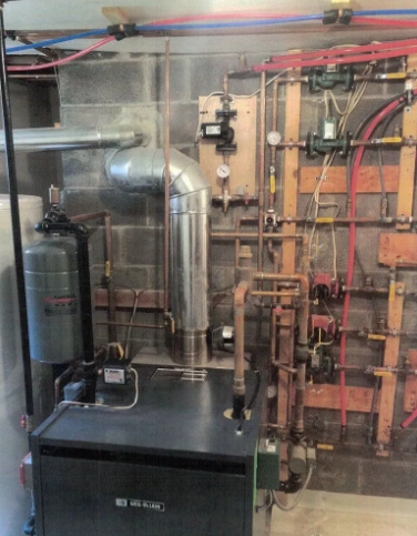 A furnace and plumbing system against cinder block wall, with copper pipes and a silver exhaust chimney.