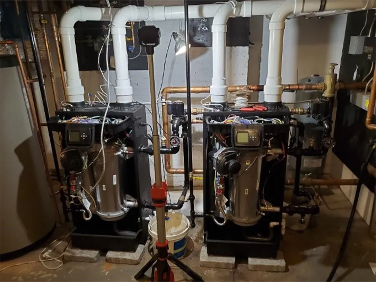 Two industrial boilers in a utility room with exposed pipes and valves behind them.