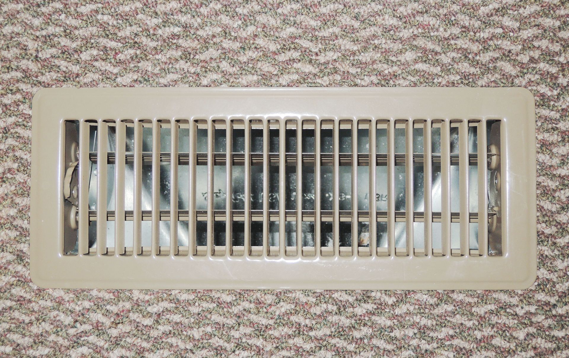 A beige rectangular floor air vent with vertical slats installed in a carpeted floor.