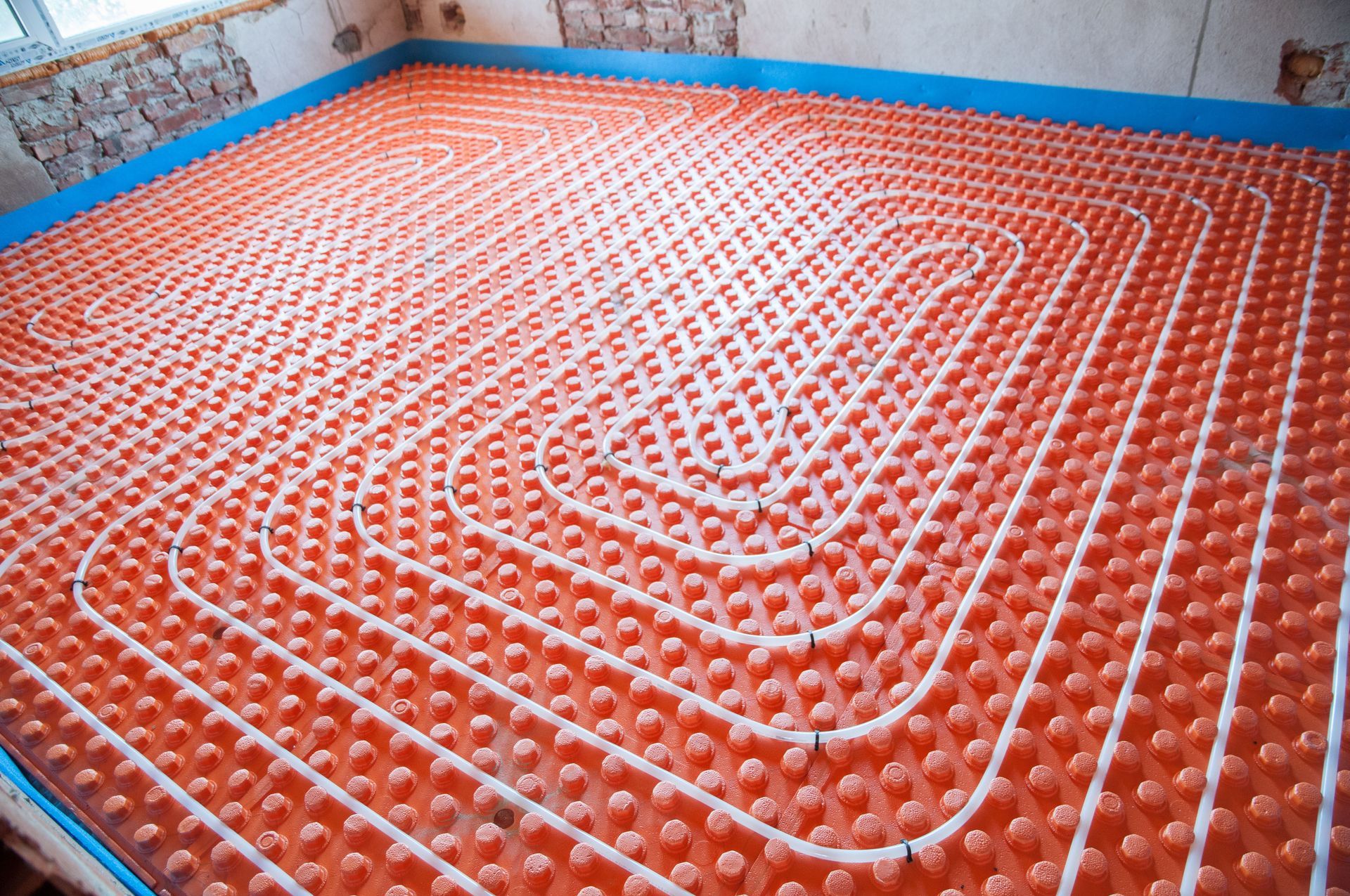 White radiant heating pipes installed in a coil pattern on orange dimpled insulation panels across a room floor.