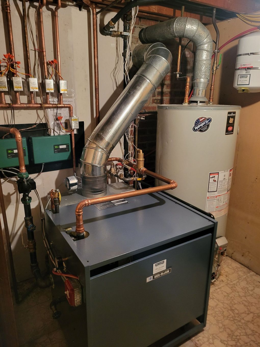 A grey heating boiler with a metal exhaust pipe next to a water heater in a mechanical room with copper plumbing pipes.
