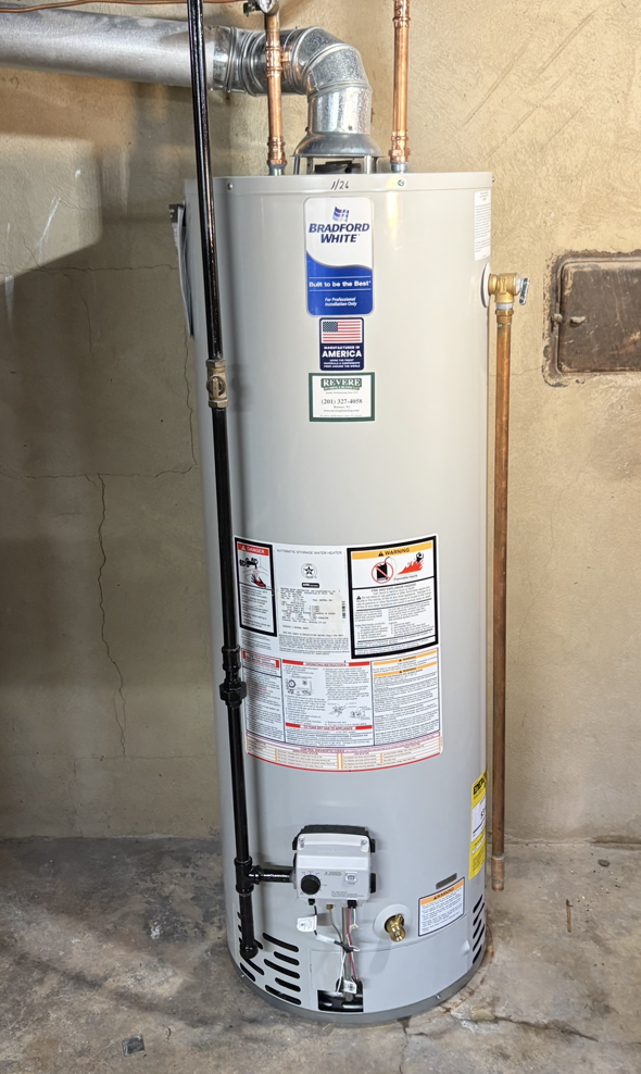 A tall, grey residential gas water heater standing against a basement wall with connected piping and vent flue.