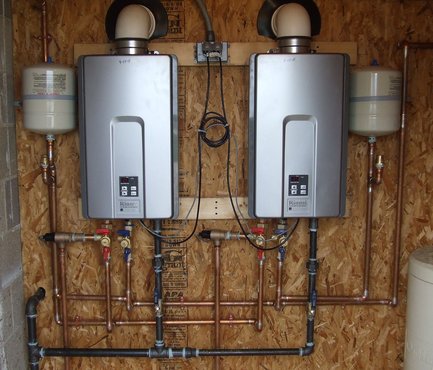Two silver tankless water heaters mounted on a wooden wall with copper piping and white expansion tanks.
