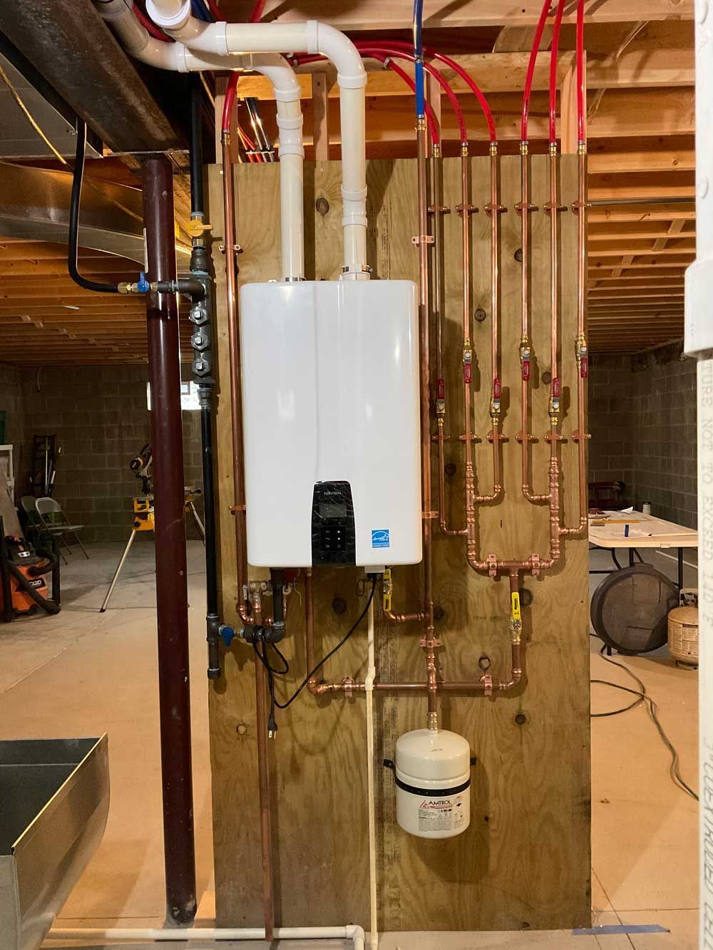 A white tankless water heater mounted on a wooden wall in a basement, surrounded by plumbing pipes and a pressure tank.