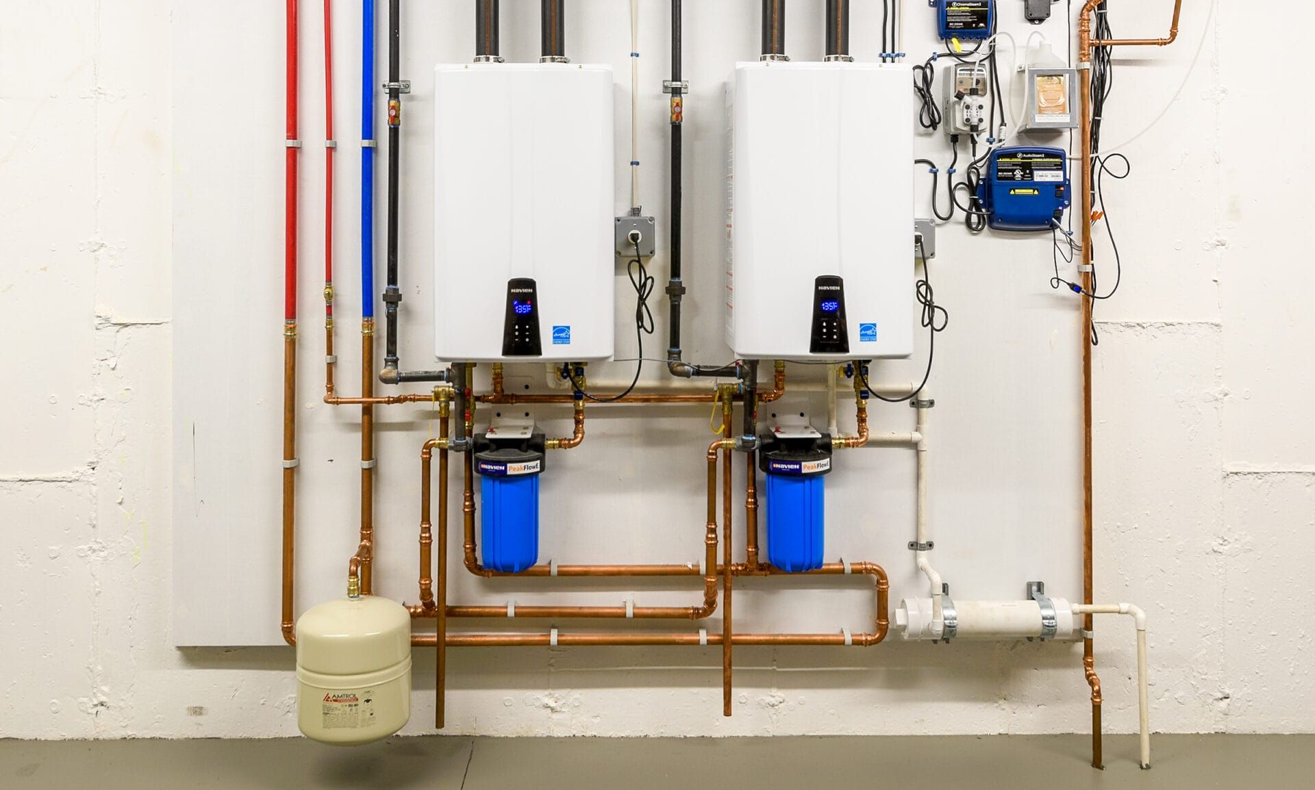 Two wall-mounted tankless water heaters with blue water filters and copper piping installed on a white wall.