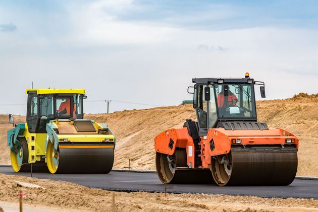 Two construction rollers, one yellow and one orange, driving on a newly paved asphalt road at a construction site.