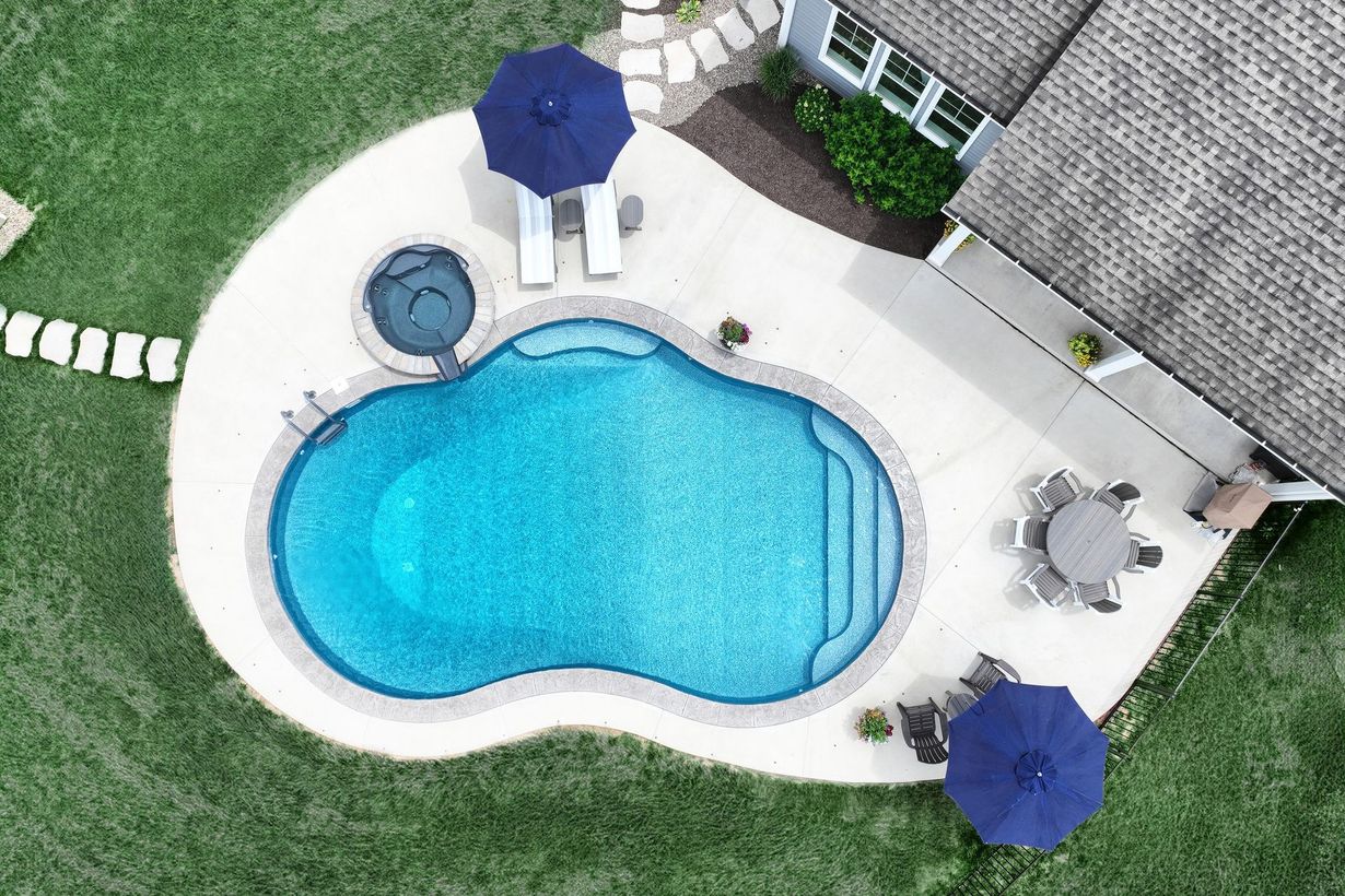 Aerial view of a swimming pool with lounge chairs, umbrellas, and a hot tub on a concrete patio next to a house.