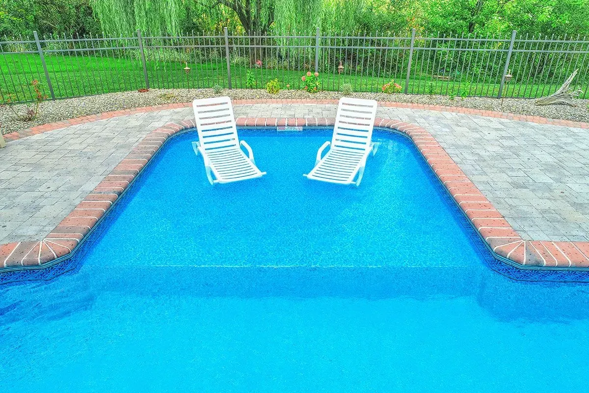 Two white lounge chairs in a blue swimming pool. Brick border. Green grass in background, fence.