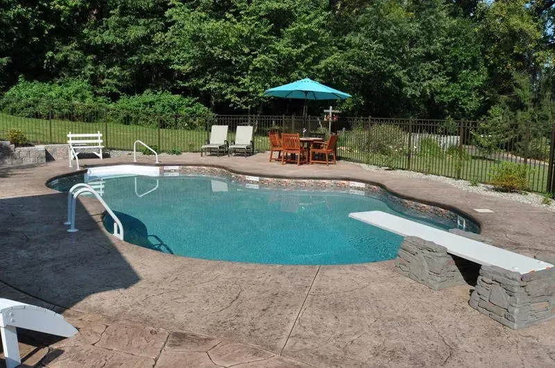 Pool with diving board, surrounded by patio, table, chairs, lounge chairs, and trees.