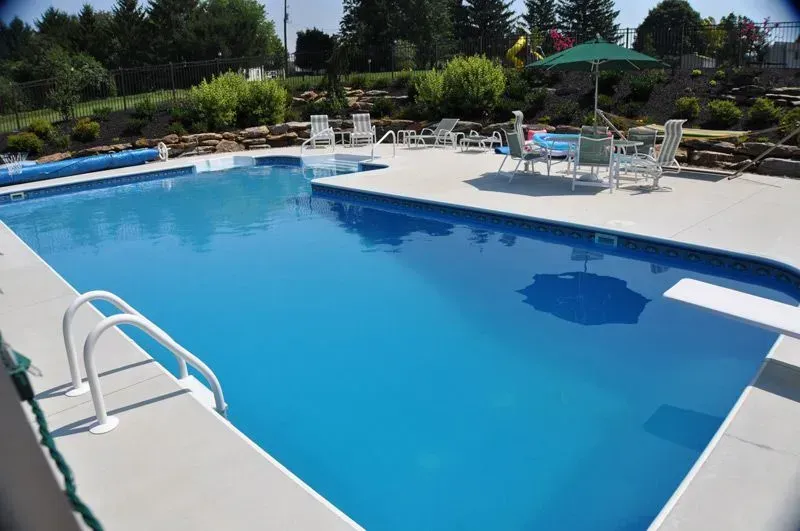 Outdoor swimming pool with blue water and white pool deck; chairs and table with umbrella on the side.