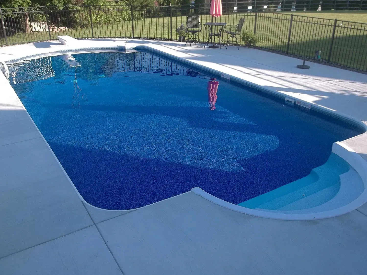 Blue-tiled swimming pool with steps, surrounded by concrete. A child floats with a pink floatie.