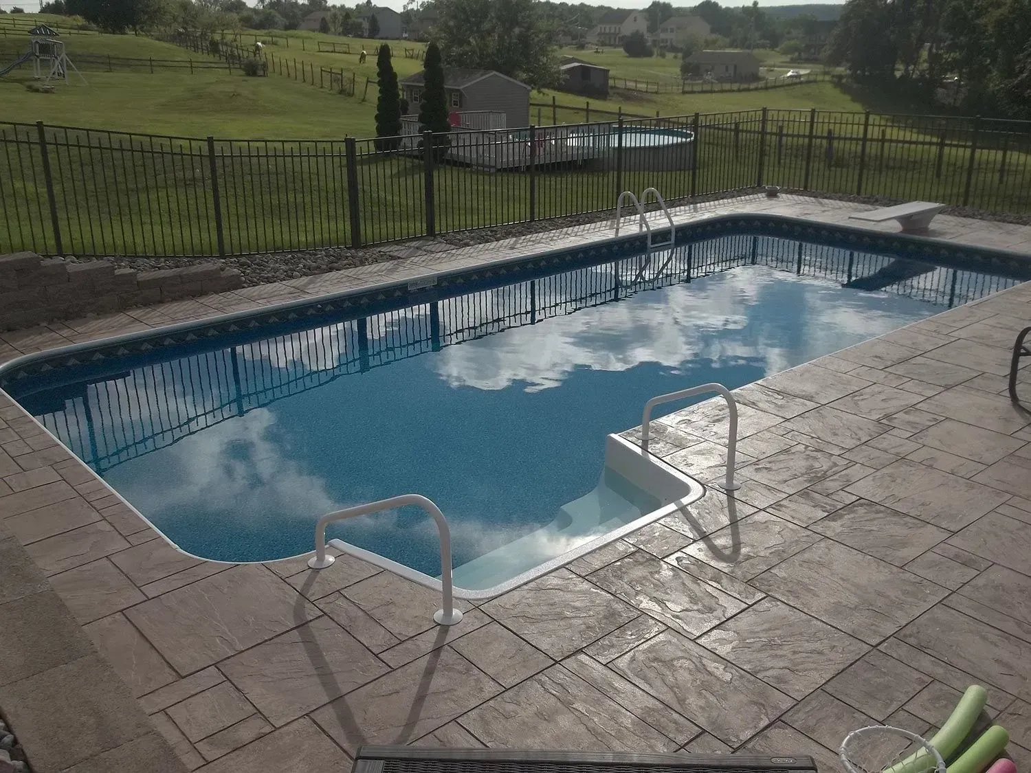Pool with sky reflection and stone patio, surrounded by black fence and green grass.