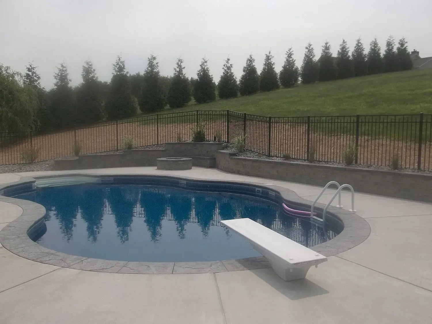 Swimming pool with a diving board, surrounded by concrete. Fenced hillside in the background.