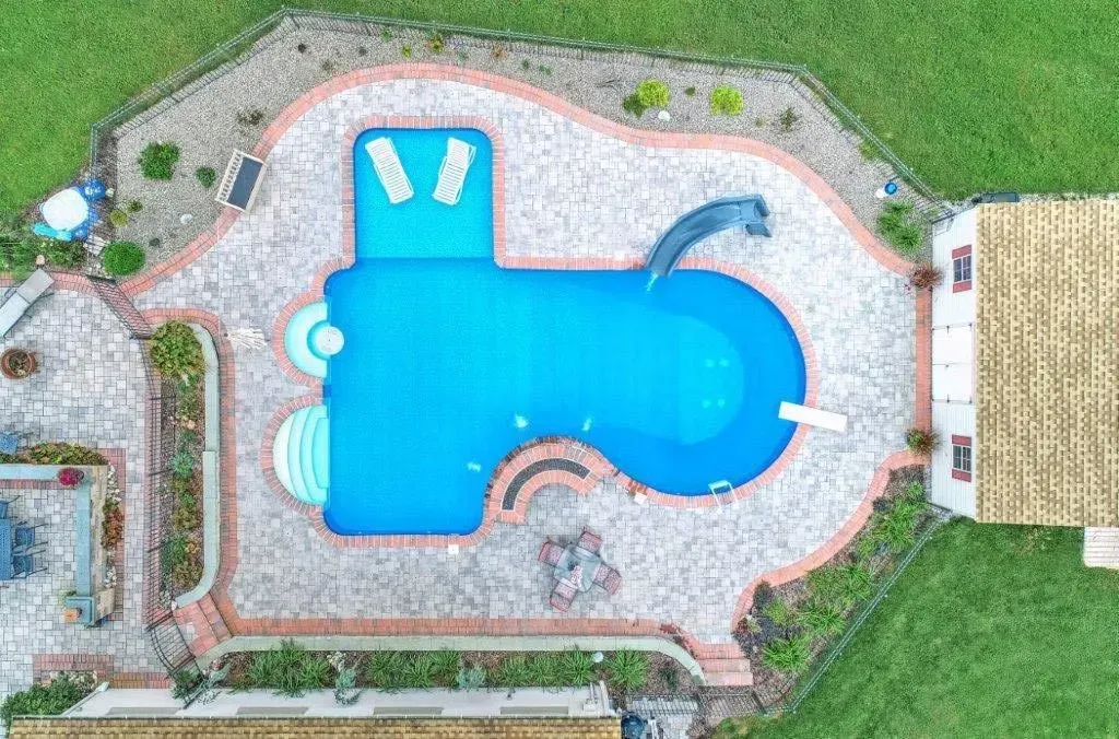 Overhead view of a guitar-shaped swimming pool with slide surrounded by a brick patio and lawn.