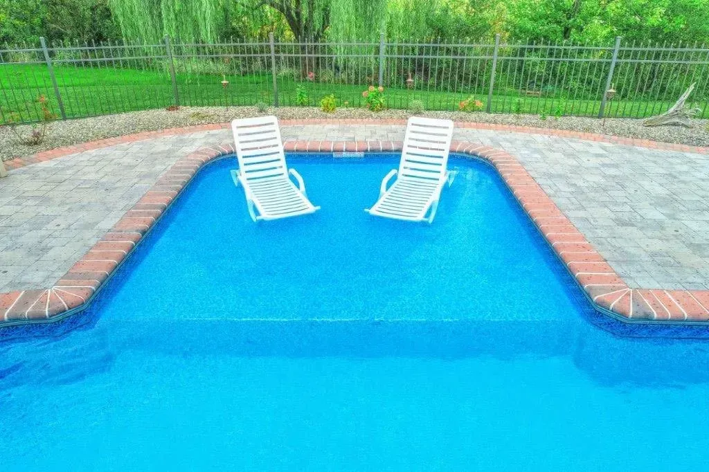 Two white lounge chairs in a shallow section of a blue swimming pool. Brick border. Green lawn behind a metal fence.