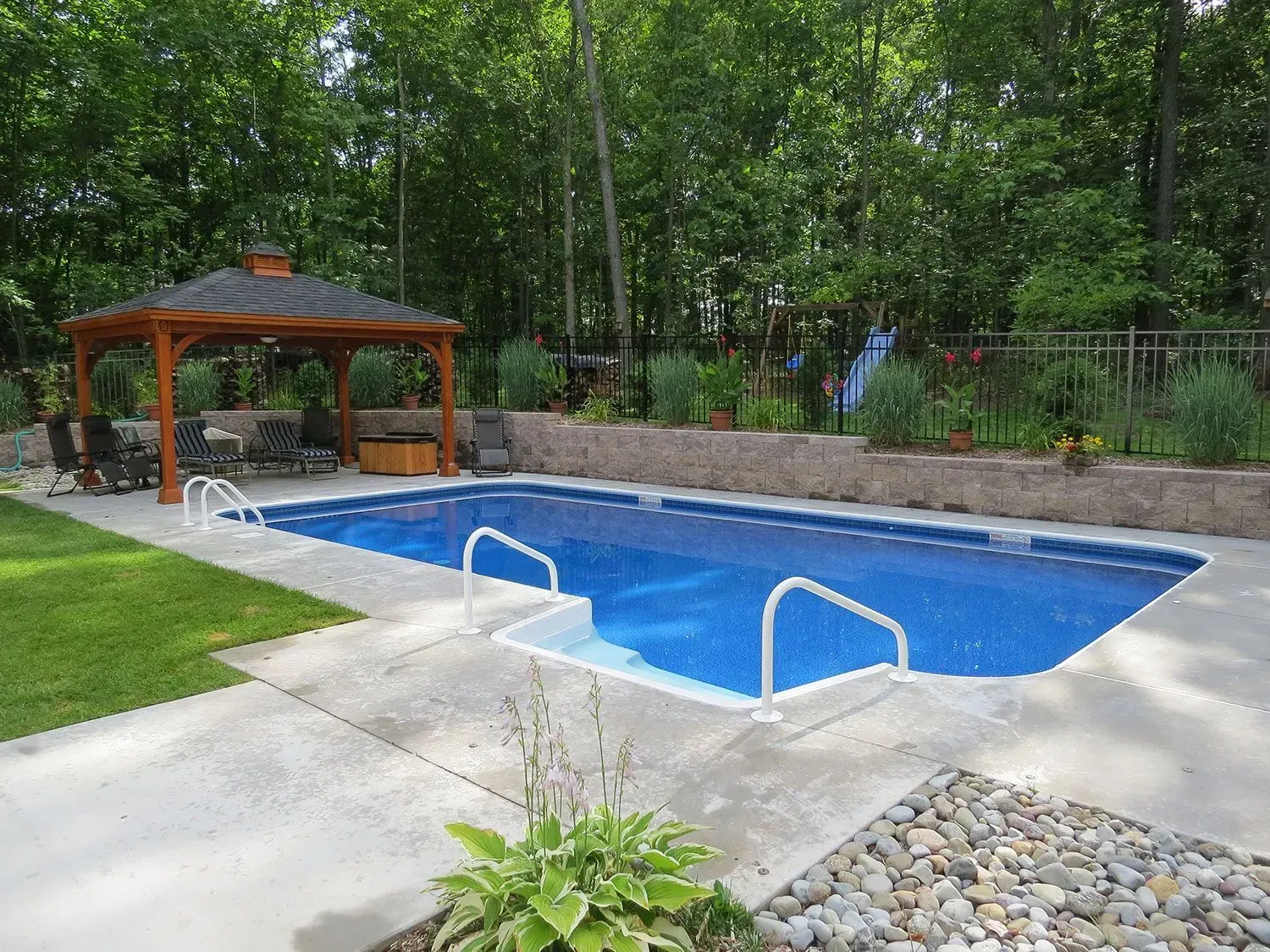 Rectangular in-ground pool with a gazebo, surrounded by concrete, lawn, and greenery.