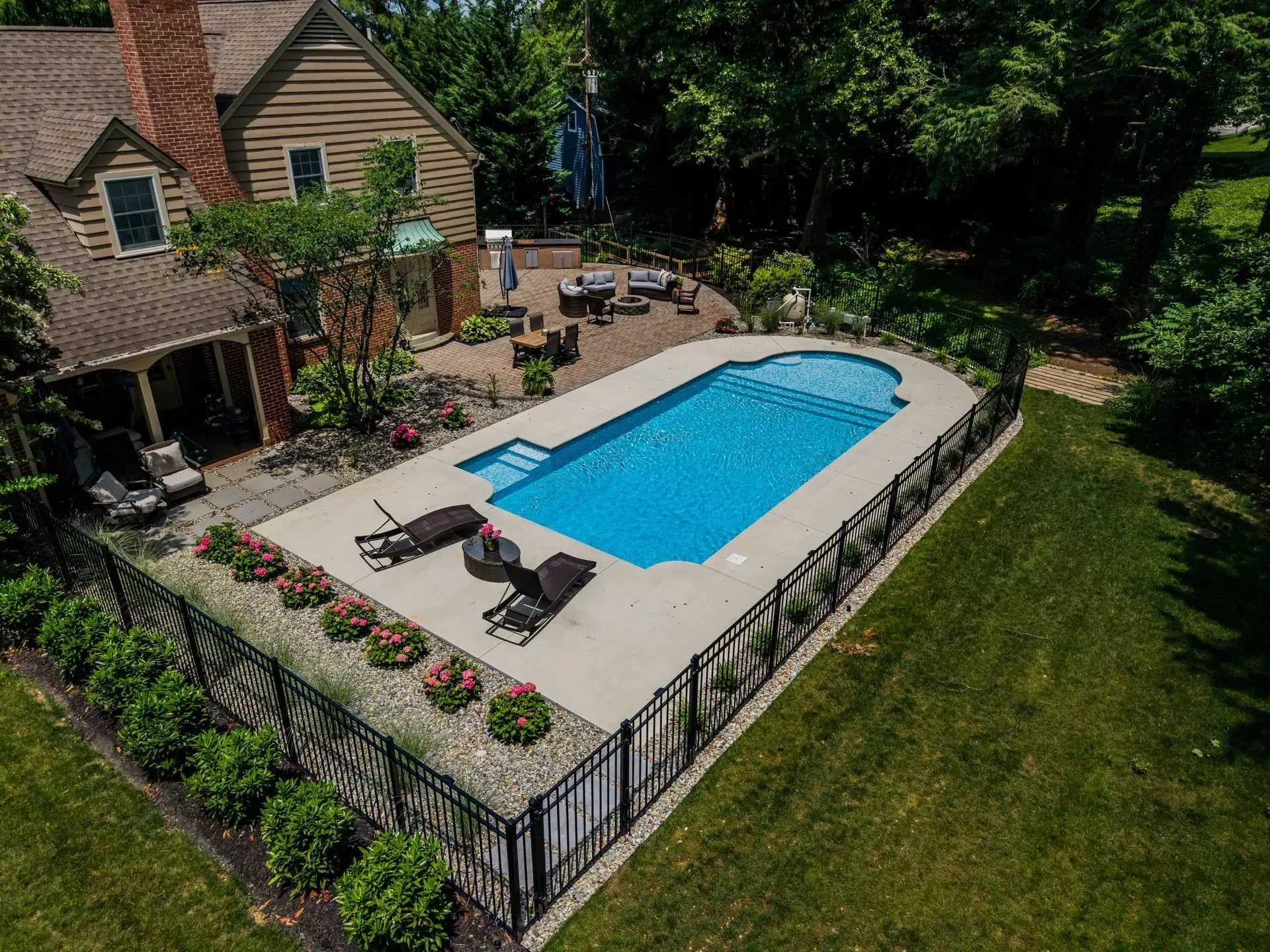 An aerial view of a backyard with a rectangular blue swimming pool, patio, and house.