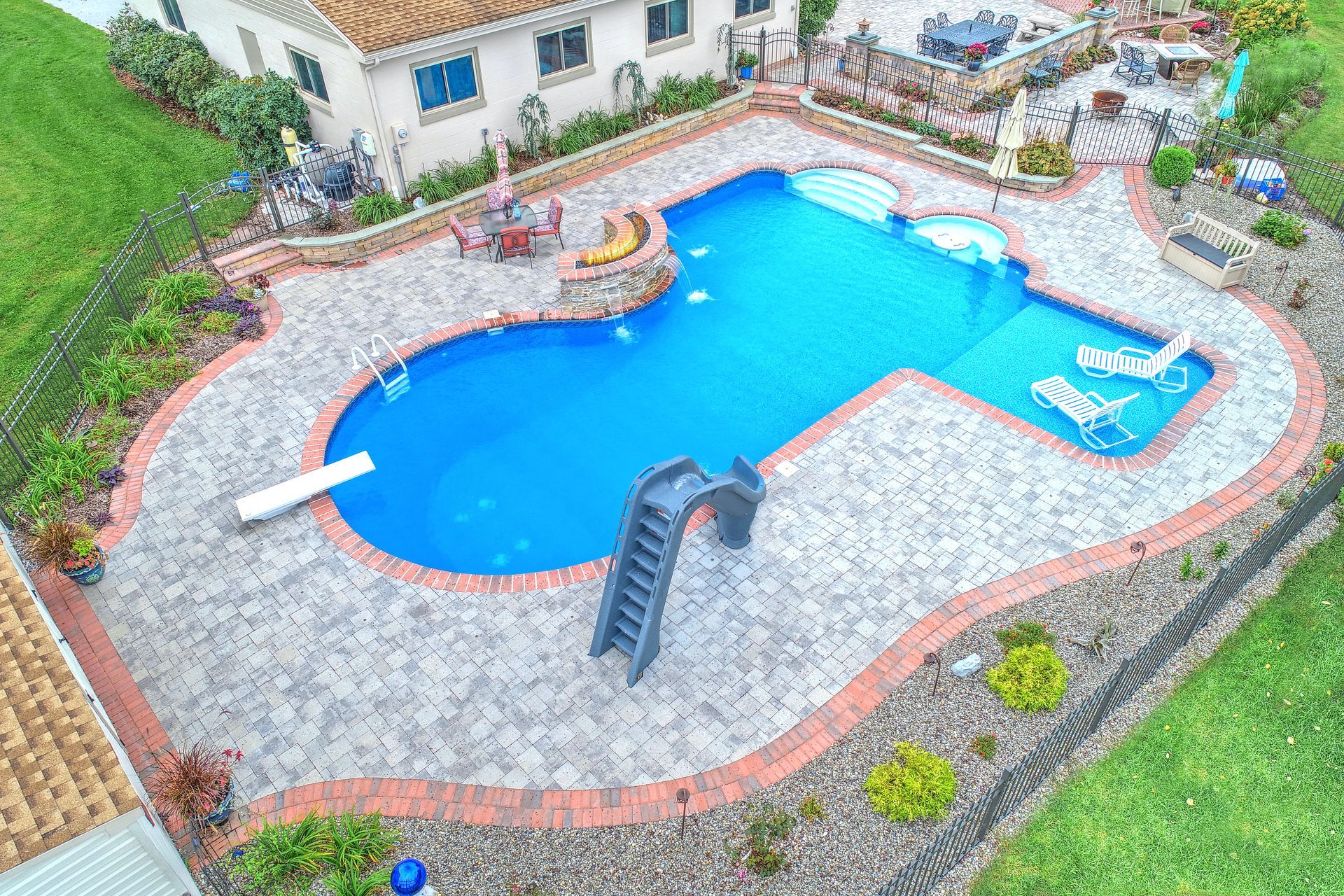 Aerial view of a custom swimming pool with a slide, diving board, and patio in a backyard setting.