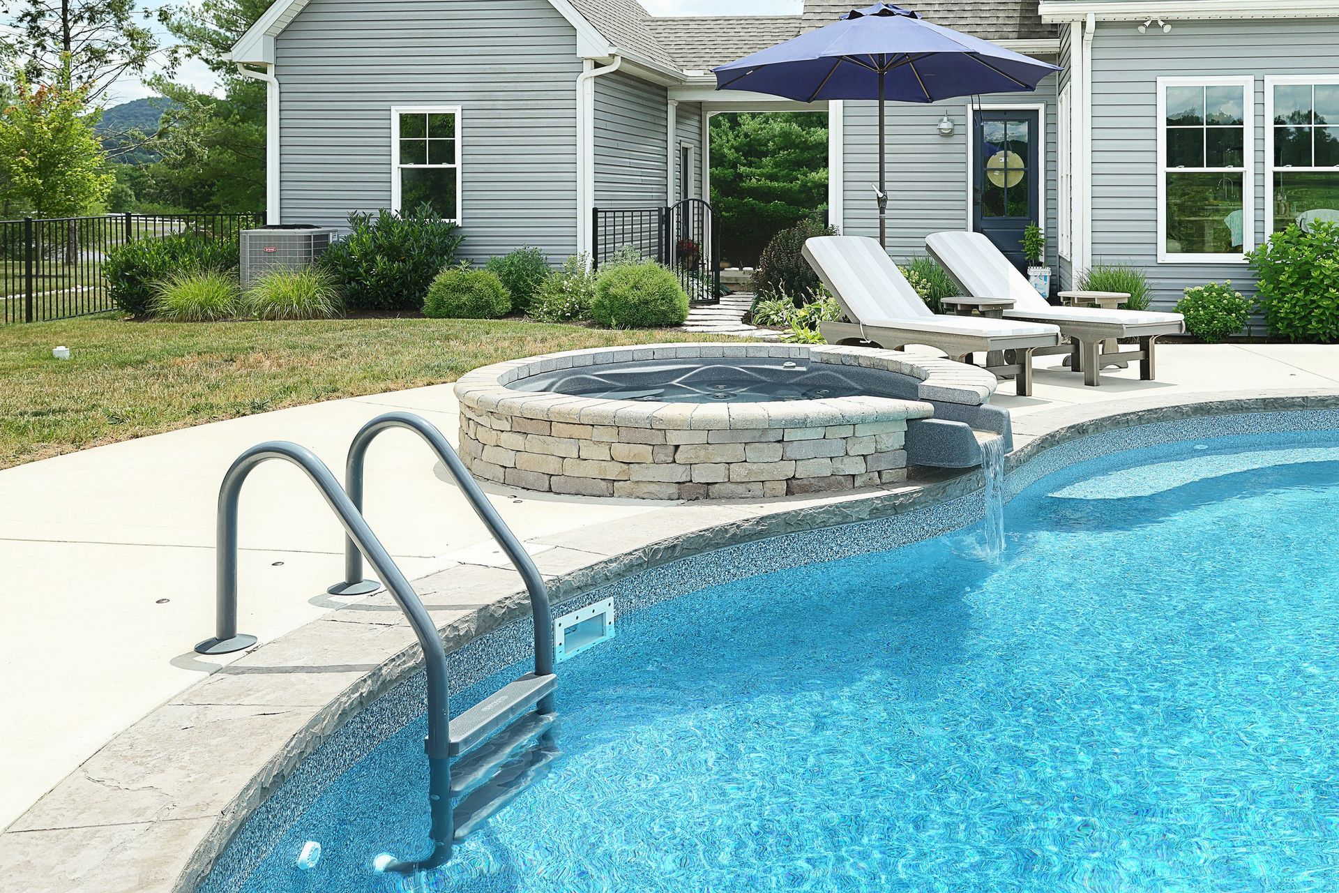Swimming pool and hot tub next to a house with lounge chairs and an umbrella.