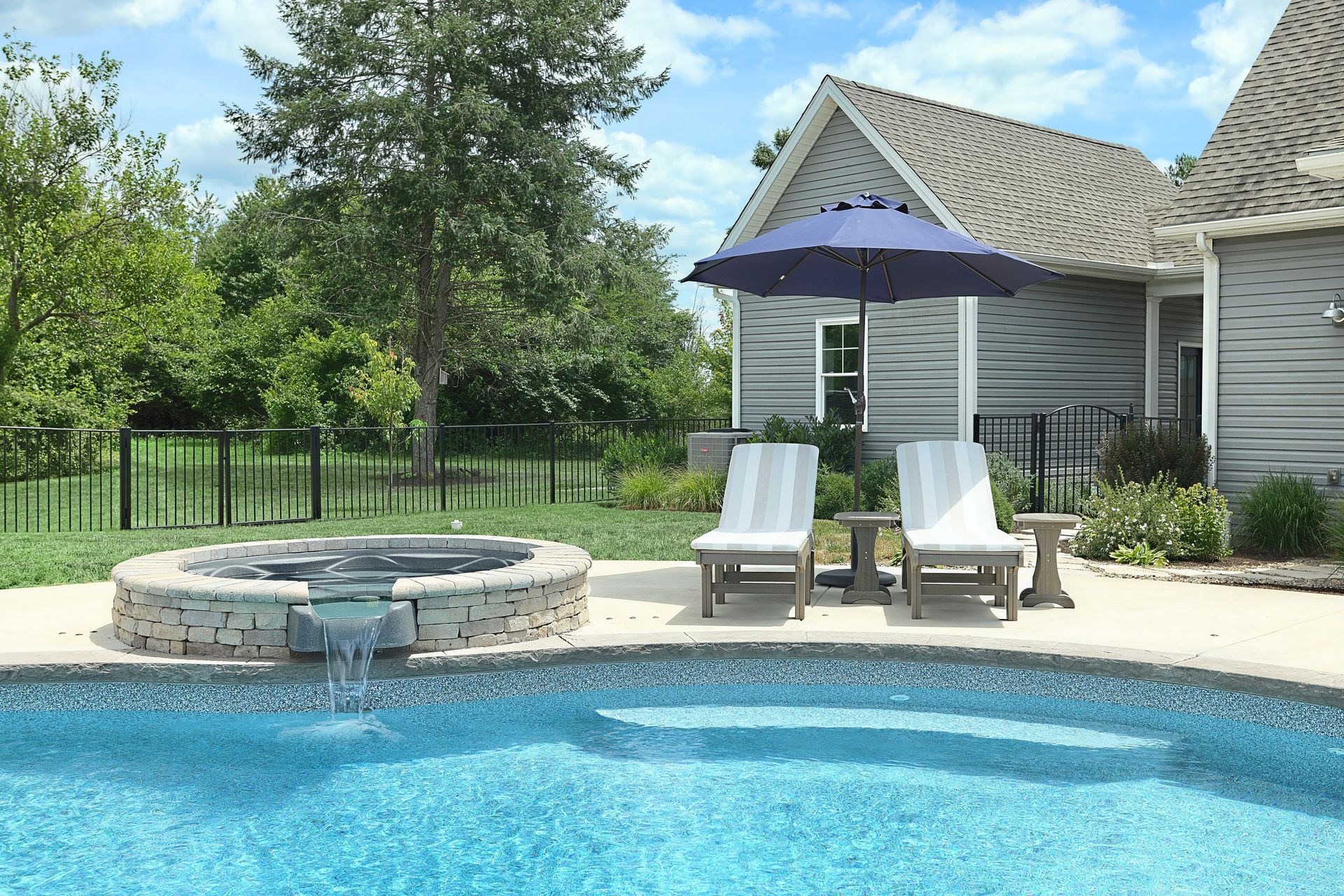 Backyard pool with jacuzzi, lounge chairs under a blue umbrella, and a gray house.