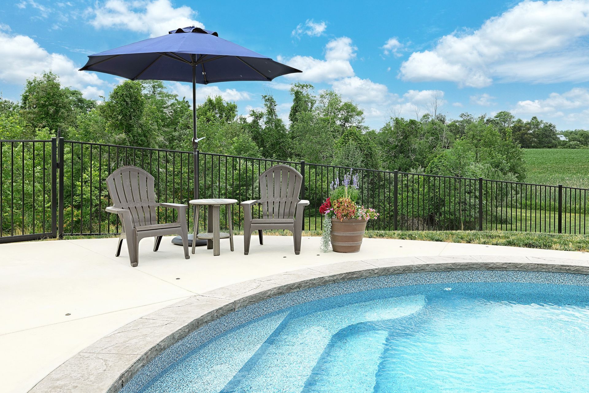 Poolside scene with chairs, table, and umbrella under blue sky.