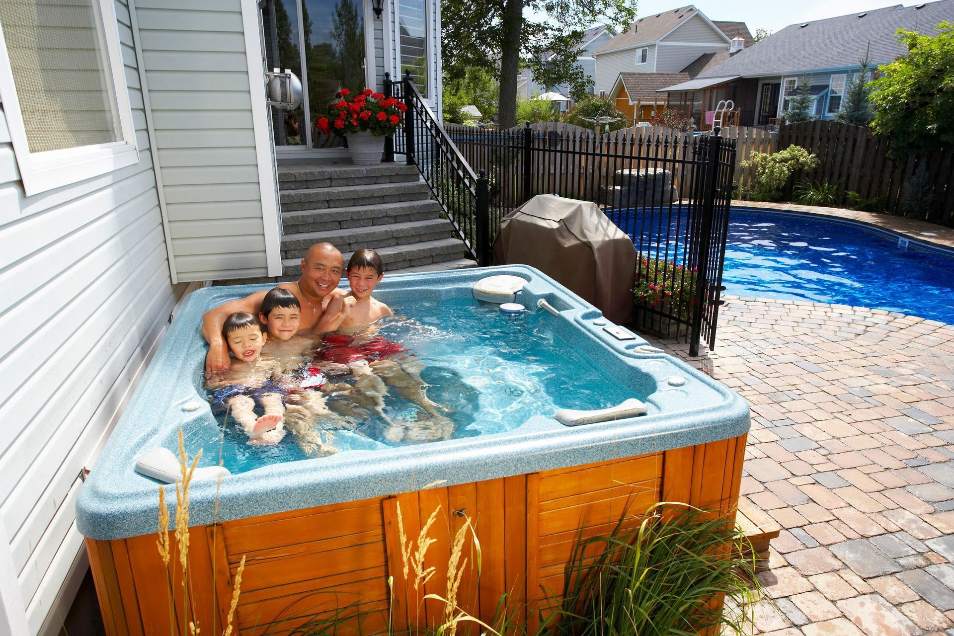 Family in a hot tub outdoors next to a pool. Wooden deck, blue water, sunny day.