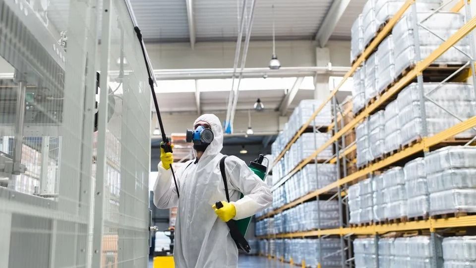 Person in protective suit spraying insecticide in a warehouse with shelving.