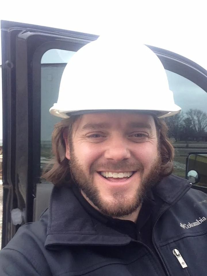 Man in white hard hat, black jacket smiling near a vehicle.