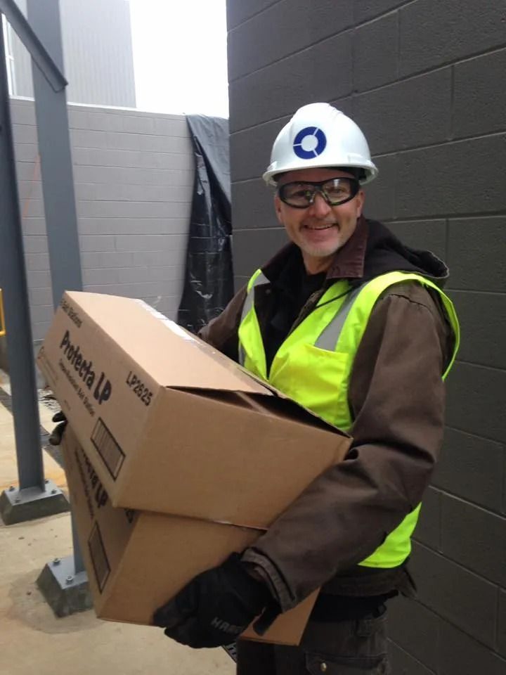 Construction worker in hard hat, safety glasses, and vest, carrying two cardboard boxes. Outdoors.