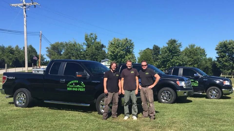 Three men stand by two black trucks in a grassy field on a sunny day. Trucks have company logo.