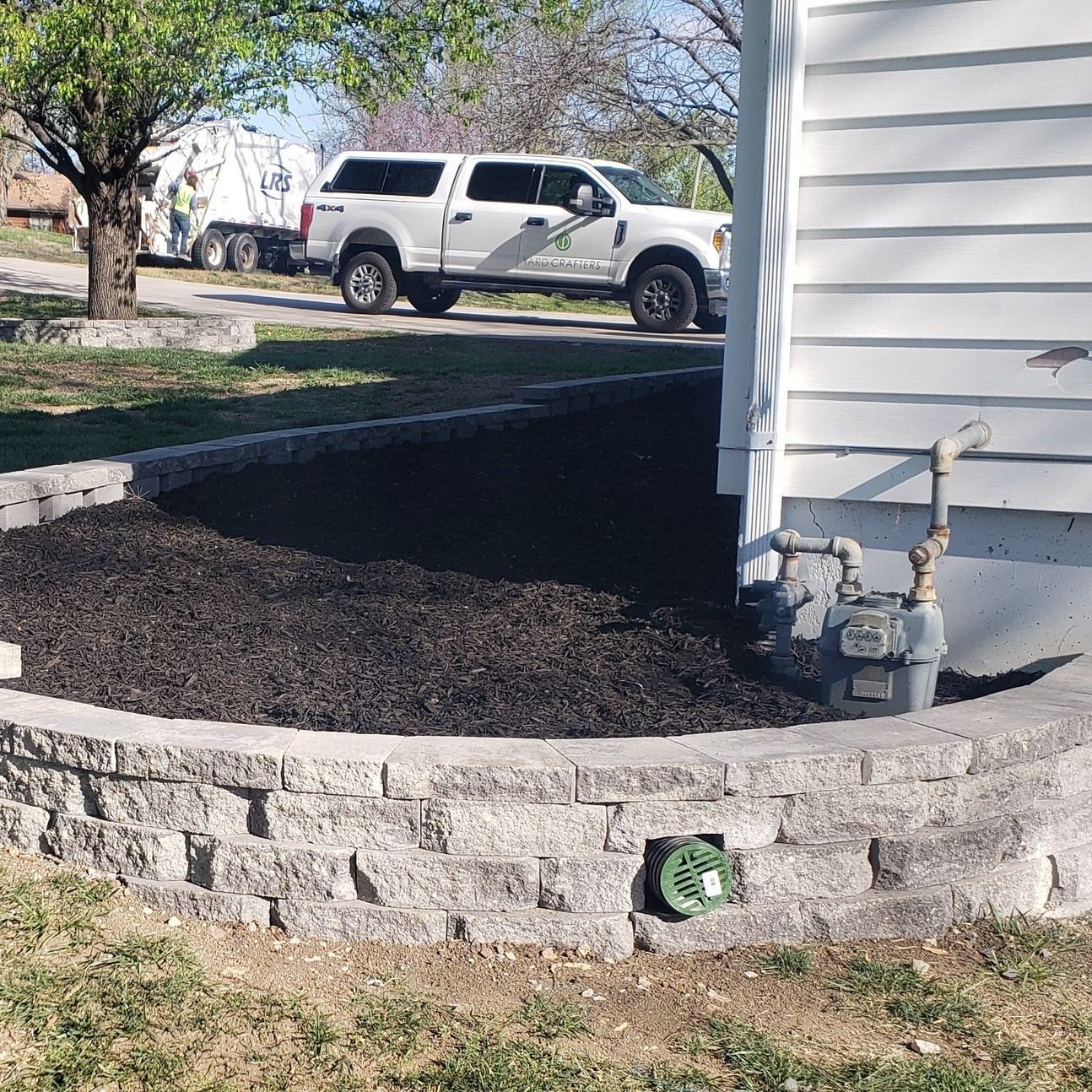 Retaining wall surrounds a mulched flower bed next to a white house, truck and tree in the background.