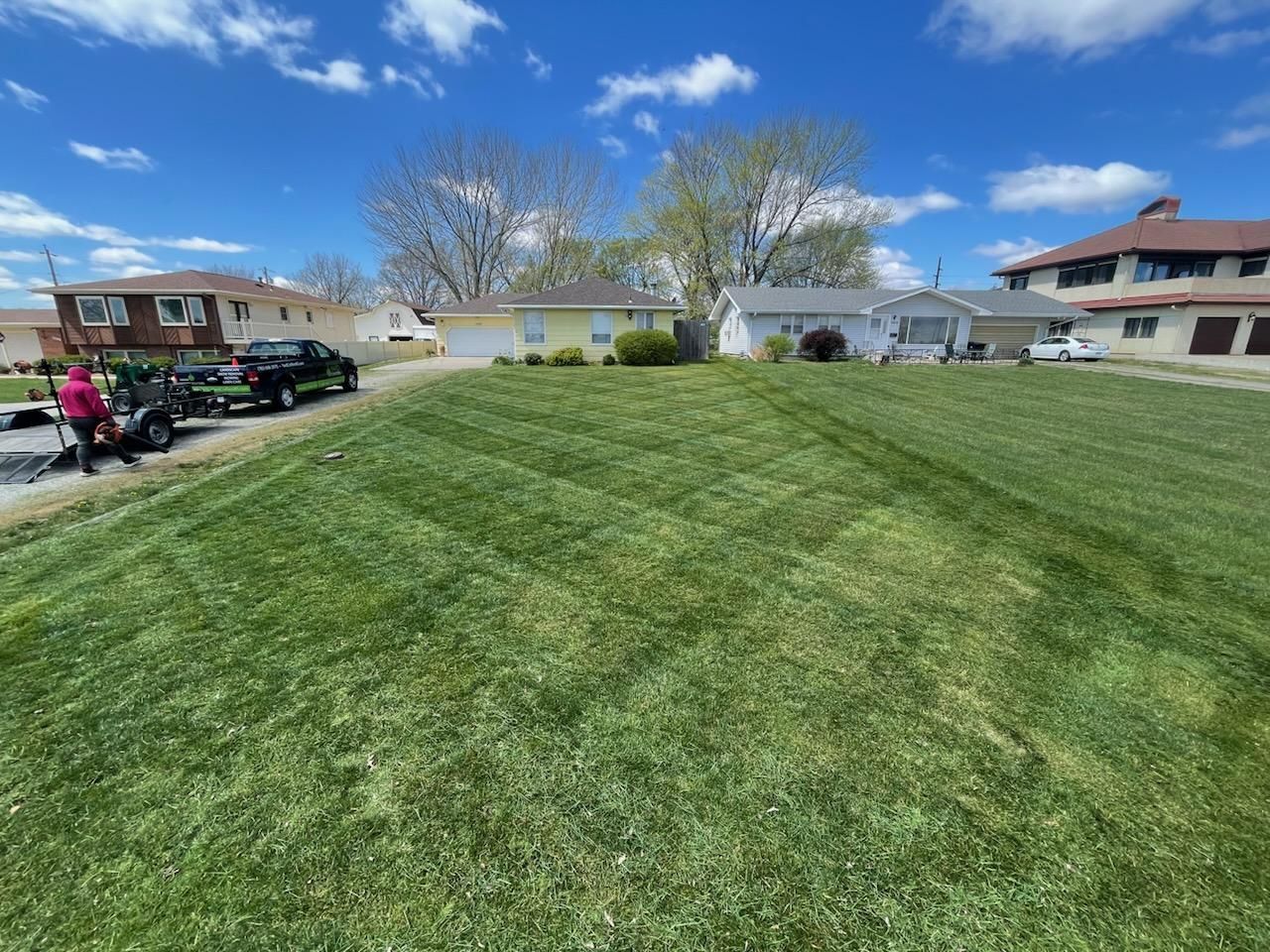 Lawn recently mowed with diagonal stripes in front of houses on a sunny day. A person and equipment are on the left.