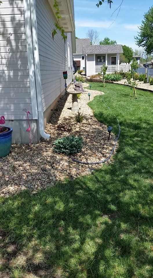 Side of a house with light siding, a gravel bed with plants, and a grassy lawn under a sunny sky.