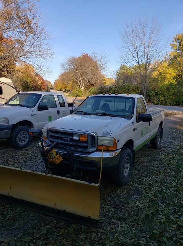 Two white trucks with green accents, parked outdoors, one with a snowplow attached.