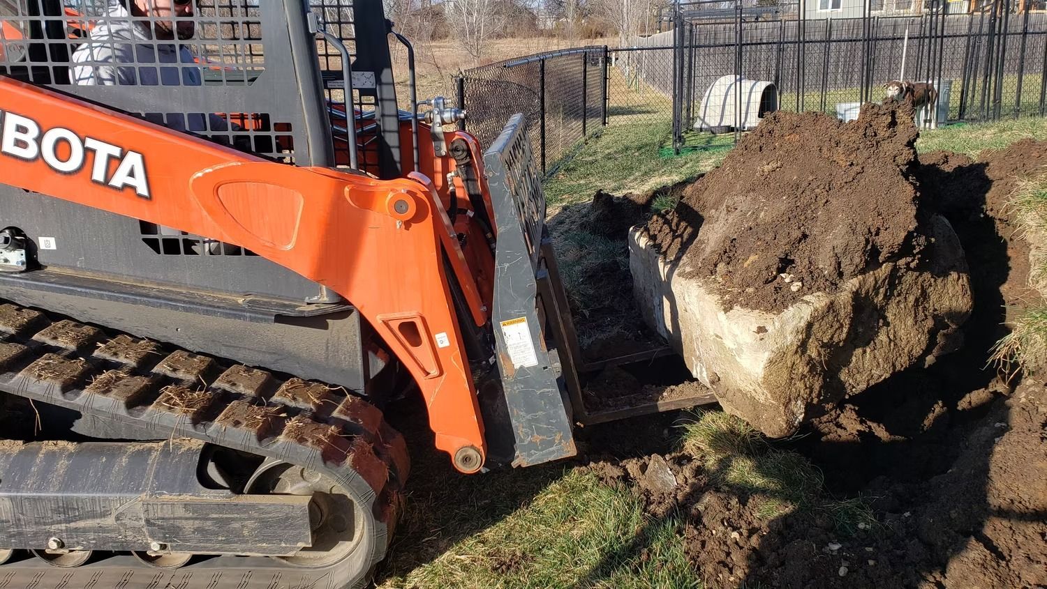 Orange Kubota skid steer lifting a large rock from a ditch, on a grassy hillside.