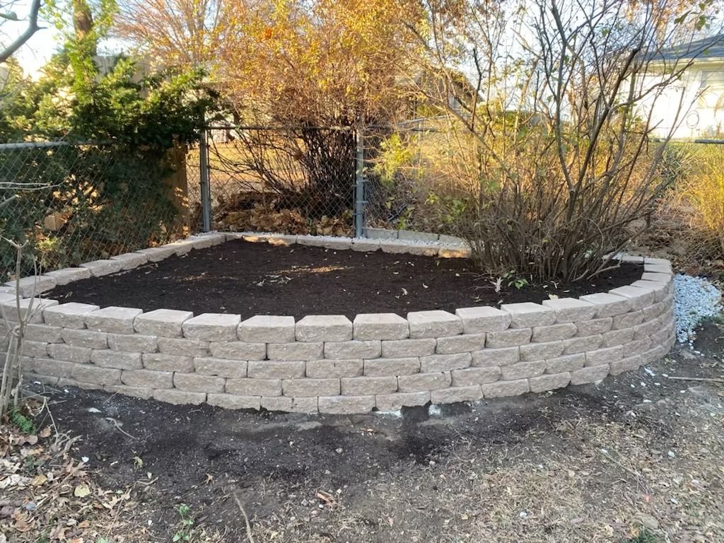 A raised garden bed constructed of stone blocks, filled with dark soil, with plants and trees in the background.