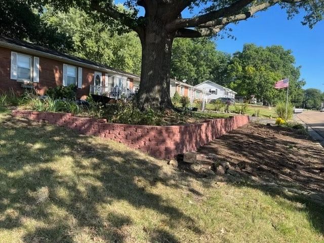 Brick house with a large tree in front. Red brick retaining wall, grassy yard, blue sky.