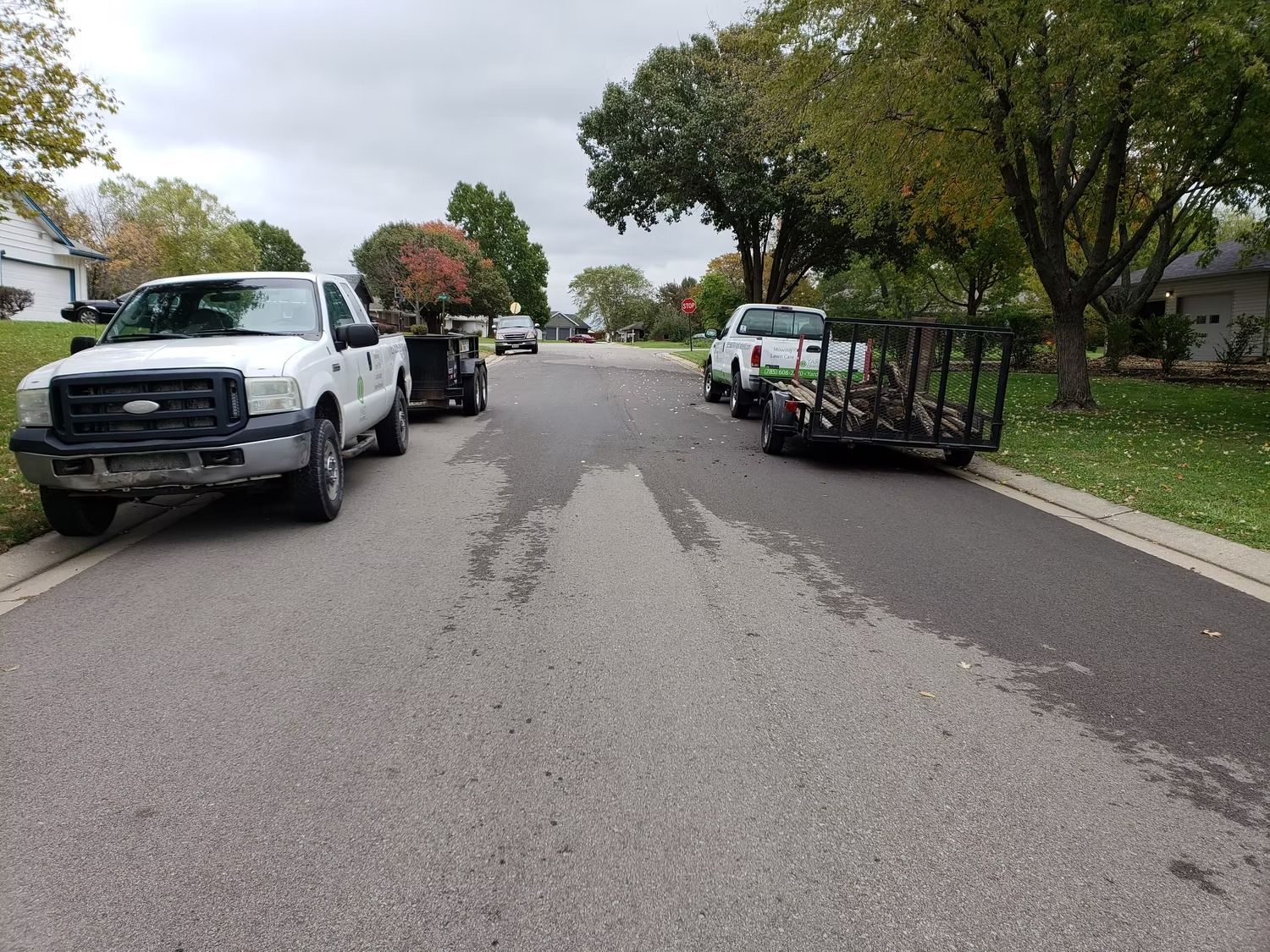 White trucks and trailers parked on a residential street with wet pavement and fall foliage.