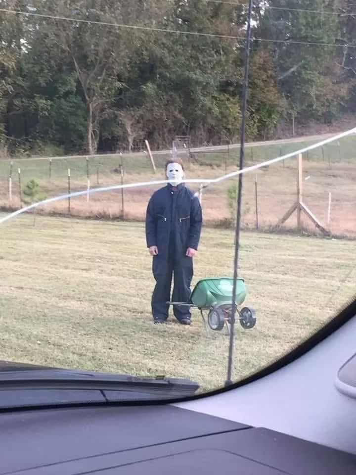 Michael Myers, in costume, stands in a yard next to a fertilizer spreader.
